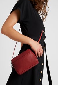 Punainen nahkainen olkalaukku suorakaiteen muotoinen, teksturoitu pinta ja säädettävä olkahihna, pidetään mustaa nappilistaista mekkoa vasten.