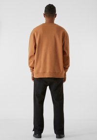 Felpa marrone con una vestibilità rilassata e polsini a costine, abbinata a pantaloni neri. La texture appare morbida, adatta per un abbigliamento casual.