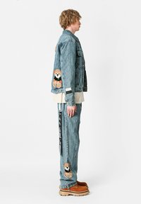 Chaqueta de denim con bordado de un oso en naranja en la parte posterior, combinada con pantalones de denim a juego que presentan un diseño similar y acentos laterales en negro.