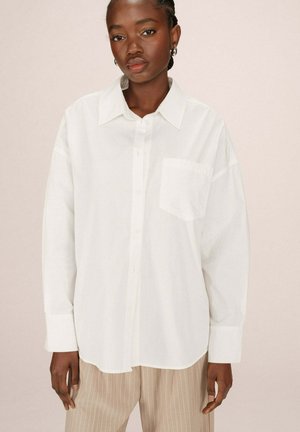 Chemise blanche à manches longues avec un col, une poche avant et une coupe décontractée, fabriquée dans un matériau doux. Associée à un pantalon beige rayé.