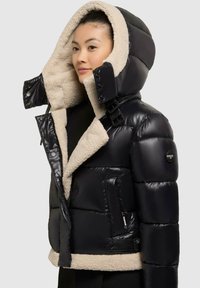 Schwarze Pufferjacke mit flauschiger beiger Innenausstattung, hohem Kragen mit übergroßer Kapuze, Knopfverschlüssen und Seitentaschen.