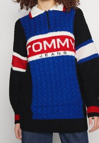 Pull en tricot bleu, noir et rouge avec un col zippé, présentant un motif en torsades et un logo "TOMMY JEANS" bien visible sur le devant.