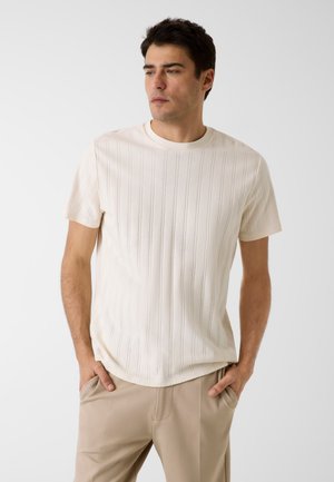 Man draagt een crèmekleurig geribd shirt met korte mouwen en beige broek, staat met handen in de zakken en kijkt opzij tegen een witte achtergrond.
