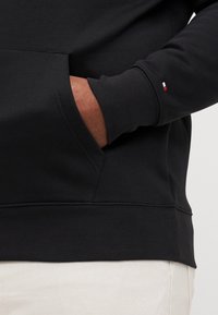 Czarna bluza z kangurzą kieszenią. Posiada ribbowane mankiety i dół. Mały trójkolorowy logo na lewym mankiecie. Miękka, teksturowana tkanina.