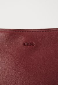 Burgundska kožna torba glatke teksture, s uzorakom otisnutog logotipa "HUGO" i preciznim šavovima duž gornjeg ruba.