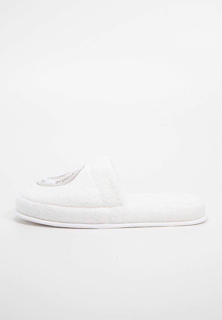 Weiße Frottier-Stoff-Slipper mit offenem Rücken und besticktem Design im oberen Bereich, flache Sohle und weiche Textur.