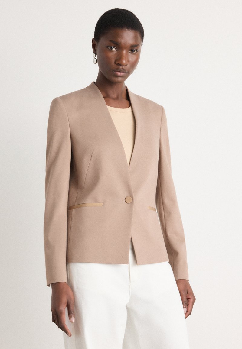 Beige tailored blazer with a notch lapel, featuring a single button closure and gold accent strips on pockets. Smooth fabric, fitted design.