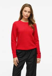 Pull en tricot texturé rouge à manches longues et col rond, associé à un pantalon noir, avec des poignets et un ourlet côtelés.
