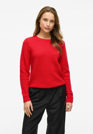 Pull en tricot texturé rouge à manches longues et col rond, associé à un pantalon noir, avec des poignets et un ourlet côtelés.