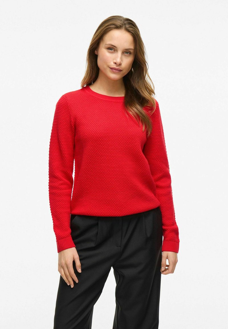 Pull en tricot texturé rouge à manches longues et col rond, associé à un pantalon noir, avec des poignets et un ourlet côtelés.