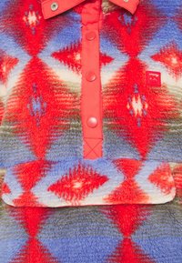 Fleece mikina s geometrickým diamantovým vzorem v červené, modré, krémové a zelené. Má zapínání na cvoček vpředu a nášivku s logem.