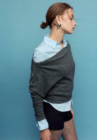 Siva, pletena majica z obliko, ki sega čez ramena, in okrasnimi srebrnimi poudarki. Nosi se preko svetlo modre srajce s črtami. Temne kratke hlače.