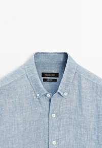 Light blue linen button-up shirt with a spread collar, small white buttons, and a textured fabric finish. Brand label visible inside the collar.