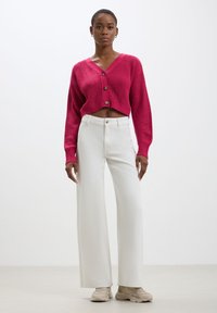Cardigan a maglia corta fucsia con texture a coste e bottoni, abbinato a pantaloni bianchi a vita alta e gamba larga, con sneakers beige.