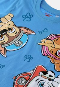 Niebieska tkanina z kolorowymi twarzami psów z serii Paw Patrol w obrożach i kaskach, otoczona odciskami łap i ikonami kości.