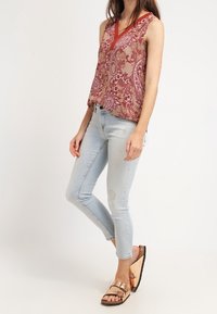 Top senza maniche in fantasia paisley rosso e crema con scollo a V, abbinato a jeans skinny blu chiaro e sandali metallici.