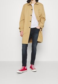 Trinchera de algodón color tan con botones negros, camiseta gris claro, jeans oscuros y zapatillas rojas de caña alta. Cuello plano, mangas largas, detalle de bolsillo.