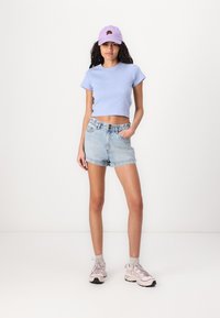 T-shirt cropped azzurro chiaro abbinato a pantaloni shorts in denim a vita alta. Il modello indossa un cappellino lavanda e sneakers rosa chiaro con calze grigie.
