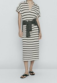 Donna che indossa un abito midi a strisce crema e nere con maniche corte, un maglione a strisce nere legato in vita e sandali neri.