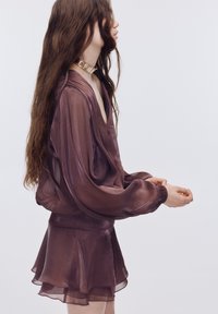 Vijolična prosojna bluza z dolgimi, volumnimi rokavi in globokim V-izrezom, kombinirana z ujemajočo se večplastno mini krilo. Zlati kovinski dodatki.