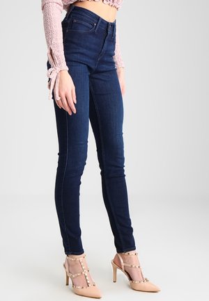 Žena má na sobě tmavě modré úzké džíny, růžový dlouhý krátký top a béžové lodičky na podpatku se sponami a cvočky.