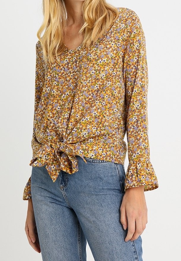 Femme portant un chemisier jaune à fleurs à manches longues avec un détail noué sur le devant et un jean bleu taille haute.
