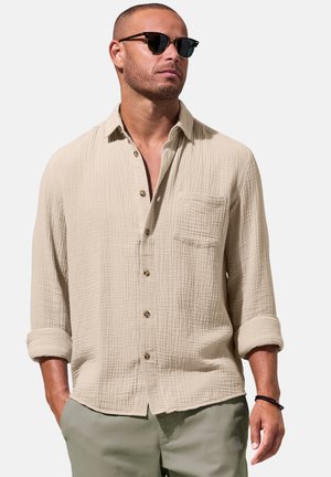 Mann trägt beige strukturierte Hemd mit Knöpfen und hochgekrempelten Ärmeln, olivgrüne Hose, Sonnenbrille und ein schwarzes Armband, steht mit den Händen in den Taschen.