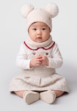 Un bébé est assis face à l’avant, vêtu d’une tenue couleur crème, d’une écharpe en tricot et d’un bonnet à pompon, les mains jointes et les jambes tendues.