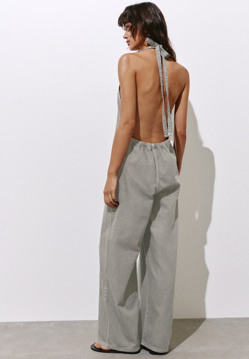 Femme aux cheveux foncés portant un haut dos nu à col halter et un pantalon gris ample taille haute, debout pieds nus contre un mur blanc.