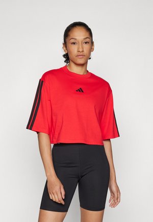 Rød kortermet t-skjorte laget av bomull med rund hals og korte ermer. Har svart tre-striper design på ermene og logo. Kombinert med svarte shorts.