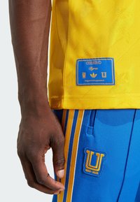 Gul sportsjersey med prægede mønstre, blå labeldetaljer, parret med blå shorts med gule striber og broderet logo.