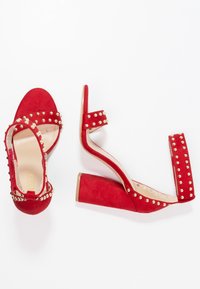 Sandalias de tacón alto de ante rojo con tachuelas doradas, diseño de punta abierta y tacón grueso. Correa para el tobillo con cierre ajustable. Forro interior suave.