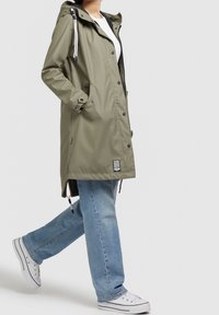Vandtæt olivengrøn parka med hætte, trykknapper, sidelommer og justerbare snore. Kombinationen med lyseblå jeans og sneakers.