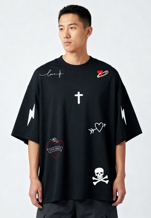 Jeune homme portant un t-shirt noir oversize avec des éclairs blancs, des cœurs, une croix, un crâne et des motifs graphiques "love hurts".