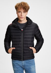 Svart puffjacka med dragkedja framtill, huva och ribbade ärmar. Har en quiltad design och två sidofickor; bärs över en grå sweater.