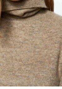 Pull col roulé marron en tissu tricoté texturé. Il présente un col montant, un matériau doux et une coupe légèrement ample.
