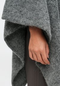 Grått filtponcho henger over en hånd, og har en myk tekstur og en asymmetrisk hemline. Tøyet ser fluffy og varmt ut.