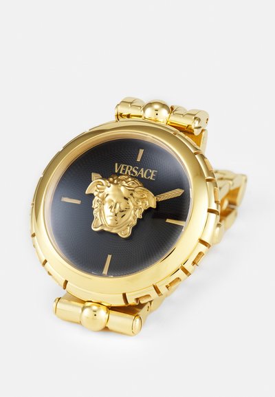 Arany színű óra fekete számlappal, középen egy kiemelkedő arany Medúza fejjel, és fém láncos karkötővel. Versace logóval ellátva.