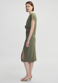 Olivengrünes Kleid mit kurzen Ärmeln, einem seitlichen Schlitz und einem gebundenen Taillendetail. Aus weichem Stoff; elegantes, knielanges Design.