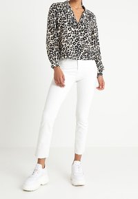 Leopardenmuster-Bluse mit lässigem Schnitt, langen Ärmeln und Knopfleiste, kombiniert mit weißen knöchellangen Hosen und robusten weißen Turnschuhen.