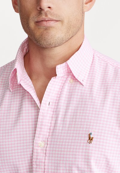 Camisa de botones a cuadros de color rosa claro con un cuello clásico y un pequeño logotipo de caballo bordado en el pecho. La tela parece tener textura.