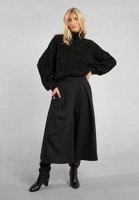 Donna bionda che indossa un maglione a collo alto nero a pois, pantaloni culotte neri a gamba larga con tasche con bottoni e stivali neri con tacco.