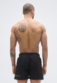 Fekete fürdőnadrág elasztikus derékpánttal, finom logó részlettel. A modellen egy tetoválás látható a háton, részletes mintákkal.