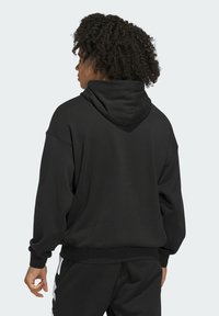 Persona con capelli ricci che indossa una felpa con cappuccio nera e pantaloni neri, rivolto di spalle su uno sfondo chiaro e uniforme.