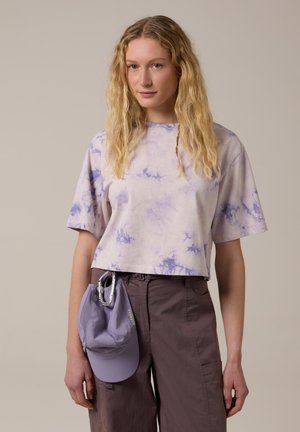 Jonge vrouw met golvend blond haar, gekleed in een paarse tie-dye shirt, bruine broek, en een paarse pet vasthoudend die aan een karabijnhaak hangt.