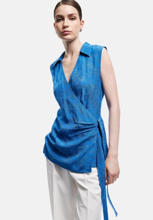 Vrouw draagt mouwloze blauwe bloemen wikkel top en witte broek, staand tegen een effen witte achtergrond.