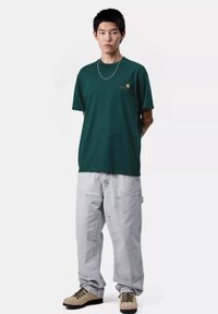 Camiseta de algodón verde oscuro, de mangas cortas con un pequeño logo, combinada con pantalones holgados gris claro y zapatos beige con cordones.