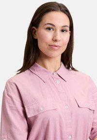 Różowa koszula z guzikami, z kołnierzykiem, z dwiema kieszeniami na piersi i białymi guzikami. Materiał wygląda na lekki o gładkiej fakturze.