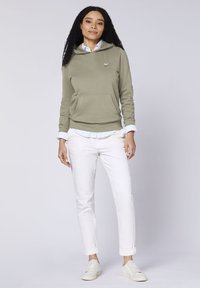 Olive green hoodie with a kangaroo pocket, layered over a light blue striped shirt. Paired with white slim pants and beige sneakers.