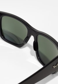 Ochelari de soare negri cu lentile verzi închise, realizați din plastic. Designul prezintă un cadru elegant și un logo proeminent pe lateral.